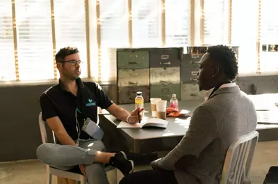 two men, one white and one black, sit across the table from one another having a conversation.