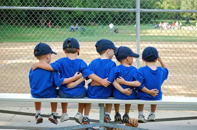 Group of five kids in matching uniforms sitting on a bench in the dugout, with their arms around each other