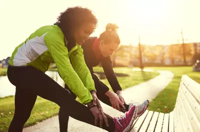 two women in running clothes stretching their legs by a park bench