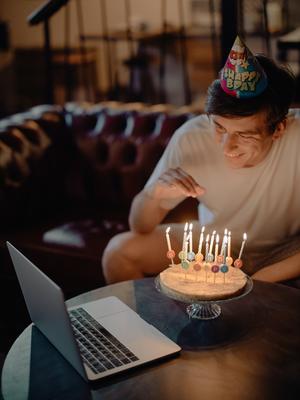 person smiling as they sit in front of a birthday cake with candles and a laptop open to a video chat