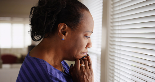 Woman stands by window with clasped hands looking worried