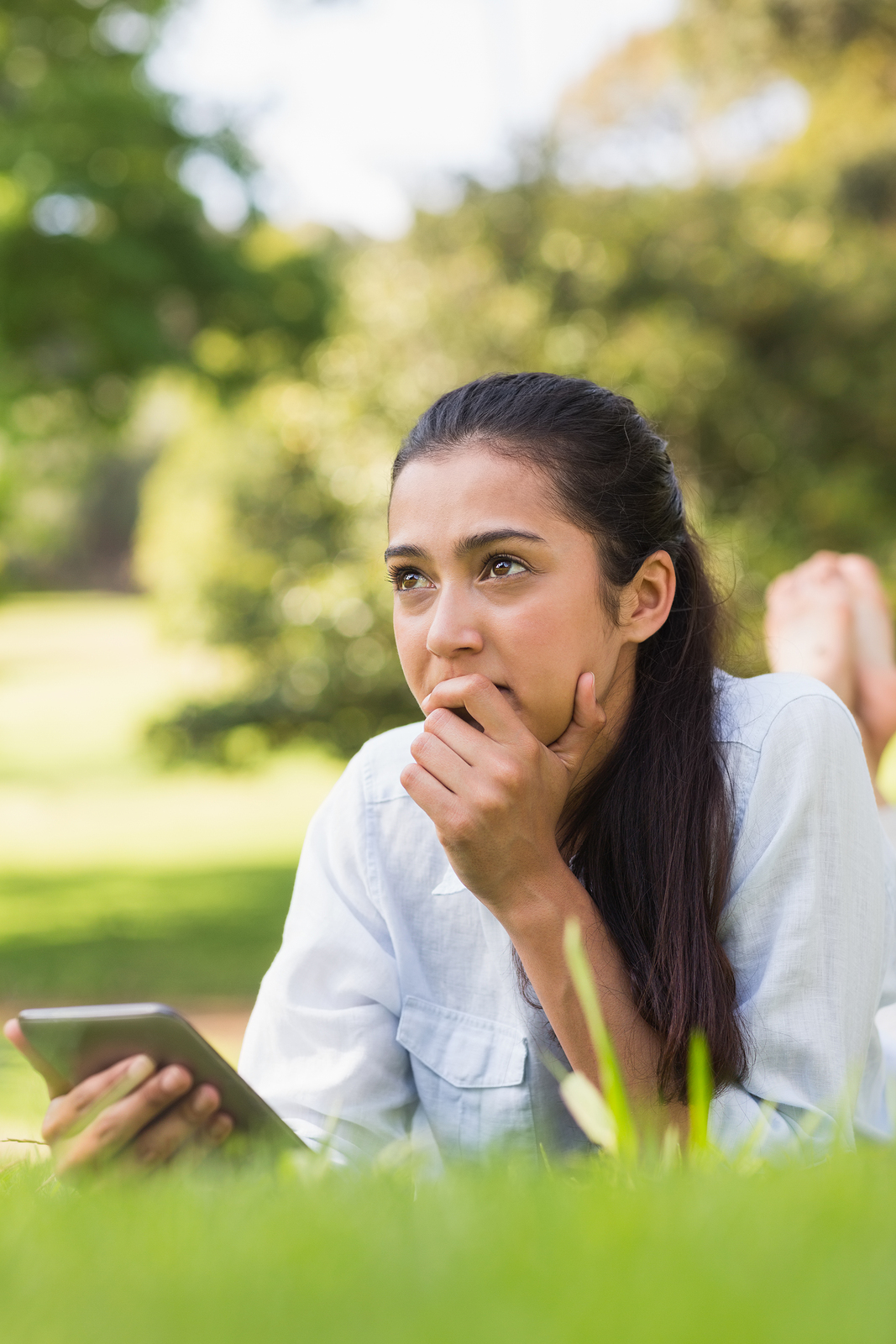 Young woman lying on her stomach in a park with a phone in her hand and a thoughtful and peaceful expression on her face.