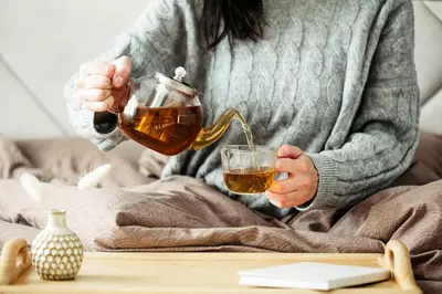 Woman in sweater pours tea into a mug while sitting in bed stock photo