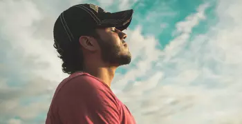 man wearing black cap with eyes closed under cloudy sky