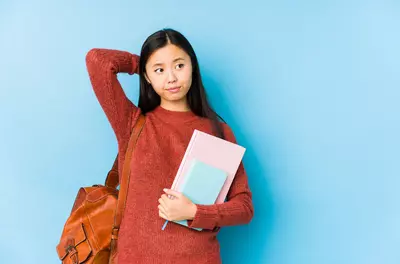 student holding books against a blue background looking pensive