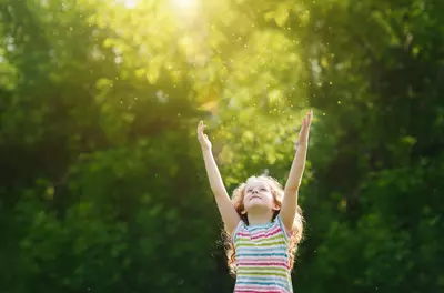 young child reaching arms up outside near green trees