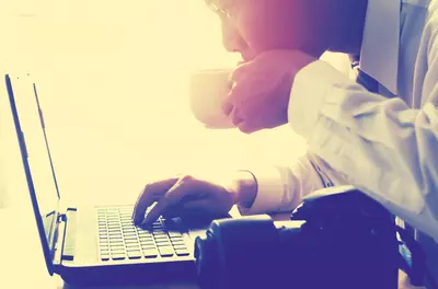 business man leaning over his laptop, reading the news while sipping coffee