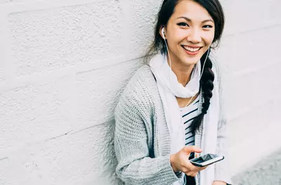 young smiling asian woman listening to a phone with earbuds while leaning against a white brick wall