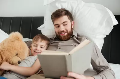 Photo of little boy lies at home in bed hugging a toy bear with his bearded father reading fairytale book.