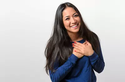 smiling woman standing against white background with hands on her heart in gratitude