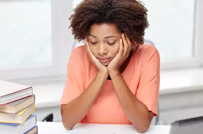 college student sitting at desk with her head in her hands next to stack of books