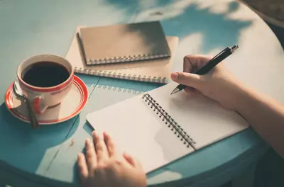 close up of hands writing in a journal, with a cup of coffee to the side