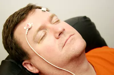 Man in orange shirt leaning back in a chair with two electrodes on his forehead