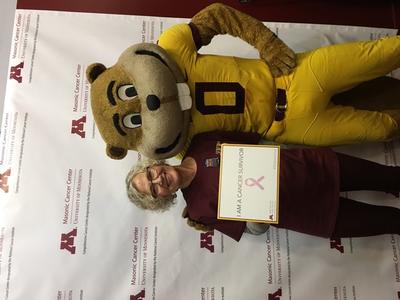 Ruth Bachman standing with U of M Mascot Golding with sign reading &quot;I&quot;m a cancer survivor&quot;.
